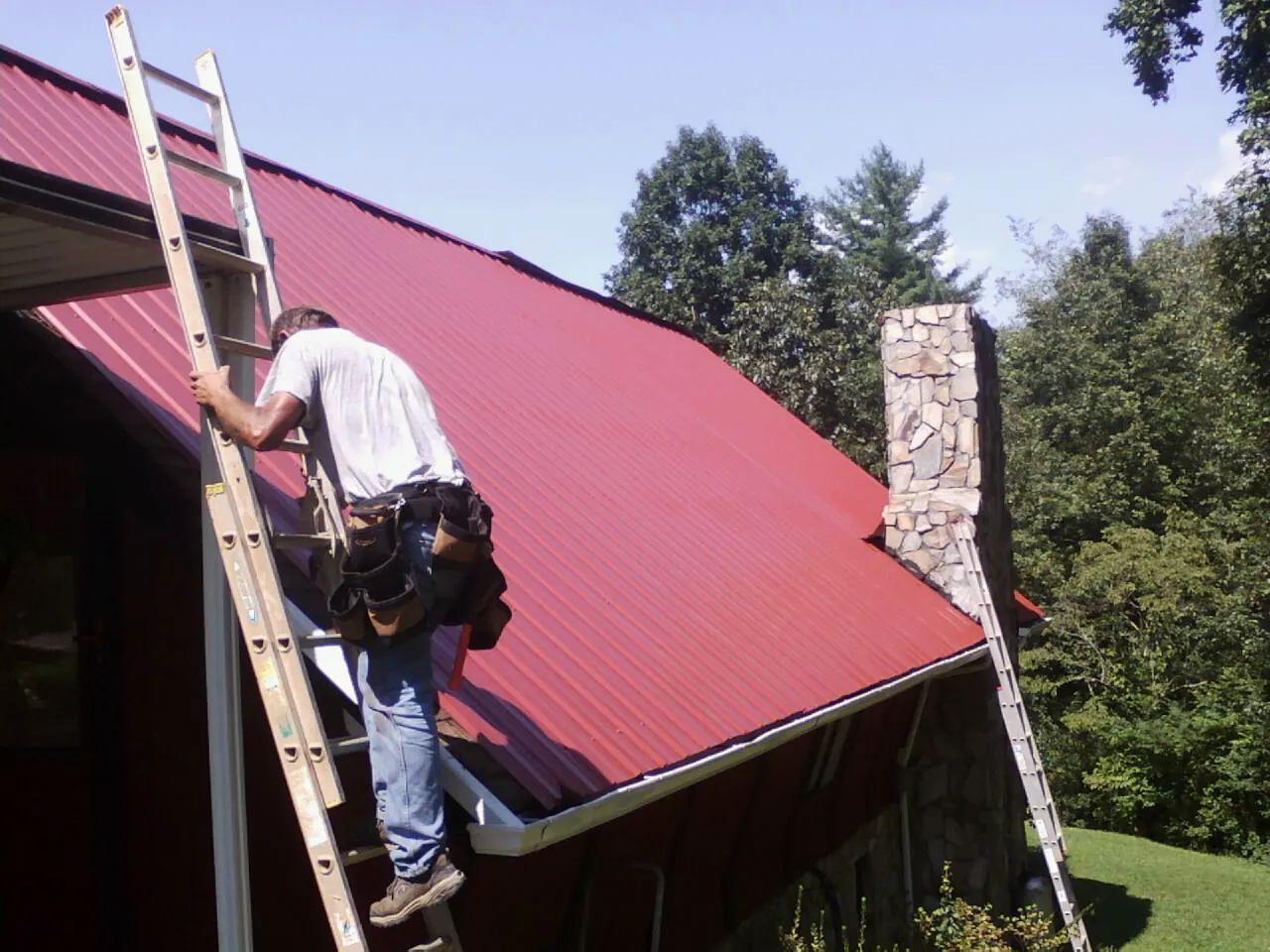 Roofer on a ladder beside a red metal roof and stone chimney — Soffit & Fascia Repair by Roofing Brothers in Terry